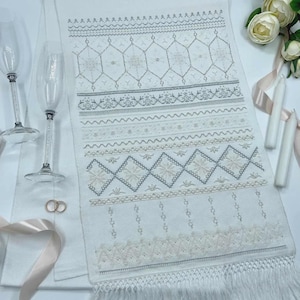 May include: A white rectangular table runner featuring intricate grey and beige embroidery in geometric and floral patterns, finished with a fringed edge. Two clear glass champagne flutes, two gold rings, two white candles, and silk ribbon.