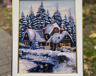 May include: A framed cross-stitch depicting a snow-covered winter scene with a cozy cabin, surrounded by snow-laden evergreen trees. The artwork is framed in white with a gold inner border, displayed on a small wooden easel.