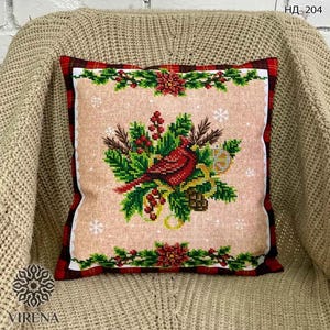 May include: A square throw pillow featuring a cross-stitch design of a red cardinal bird perched among green pine branches, red berries, and pinecones on a light tan background, framed by a red and black plaid border with poinsettia accents.