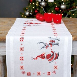 May include: White Christmas table runner with a cross-stitch design featuring Santa Claus riding a reindeer. The runner has a red and white color scheme with snowflake and geometric patterns. The runner is on a wooden table.