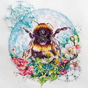 May include: A cross-stitch embroidery featuring a central bumblebee with black, yellow, and orange fur, surrounded by pink and white flowers, green foliage, and light blue circular patterns on a white fabric background with decorative bead accents.