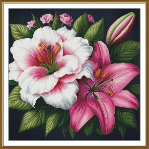 May include: Cross-stitch floral art featuring two lilies in full bloom, one white with pink accents and the other a vibrant pink. Green leaves and buds surround the flowers, set against a dark background, all within a gold frame.