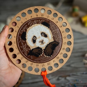 May include: A wooden thread organizer with 28 holes numbered 1 through 28. The organizer has a brown leather circle in the center with an embroidered panda bear design. The panda bear is white with black eyes and ears, and a brown nose. The organizer has a red tassel.