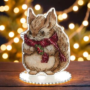 May include: A brown and white cross-stitch of a squirrel wearing a red scarf and holding a sprig of holly. The squirrel is standing on a clear acrylic base.