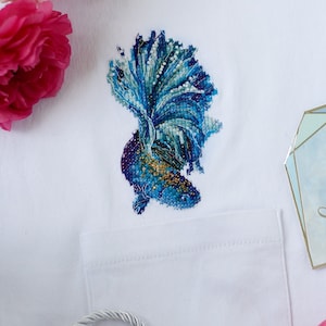 Op de afbeelding: Een wit T-shirt met een geborduurd ontwerp van een blauwe en groene vis. De vis heeft een lange, vloeiende staart en bevindt zich aan de linkerkant van het shirt.