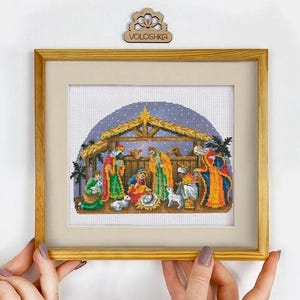 Könnte beinhalten: Ein gerahmtes Kreuzstichbild, das eine Krippenszene mit Figuren in farbenfrohen Gewändern unter einem hölzernen Stall zeigt. Ein Stern leuchtet über dem Dach und Tiere sind abgebildet. Der Holzrahmen ist hellbraun und der Markenname VOLOSHKA ist auf einem kleinen Holzschild oben zu sehen.