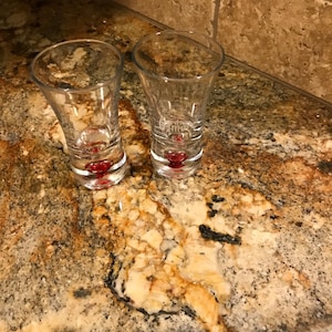 Puede incluir: Dos vasos de chupito transparentes con base roja están colocados sobre una encimera moteada. Los vasos tienen una parte superior acampanada y una pequeña base roja redonda. La encimera tiene una mezcla de tonos marrones, beiges y negros.