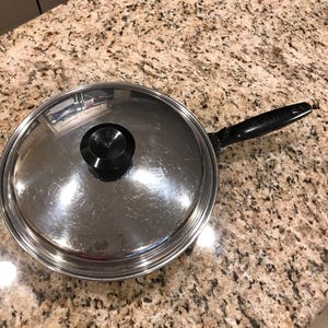 Puede incluir: Una sartén de acero inoxidable con tapa de cristal y mango negro. La sartén es redonda y tiene una superficie brillante y reflectante. La tapa tiene un pomo negro en el centro.