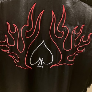 May include: Black garment with red flame embroidery and a white spade design. The flames are outlined in red thread, creating a bold contrast against the black fabric. The spade is centered below the flames.