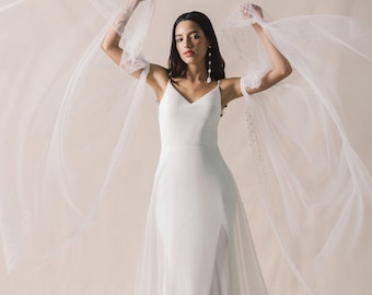 May include: A white wedding dress with a fitted bodice and a flowing skirt. The dress has a deep V-neckline and a slit in the skirt. The bride is wearing a veil with a delicate pattern of small dots.