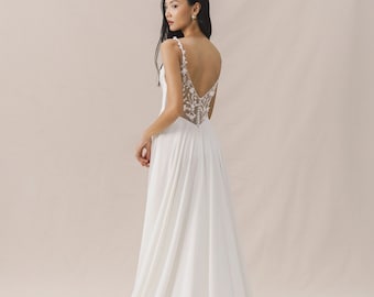 May include: A white wedding dress with a low back and a delicate floral lace design. The dress has a flowing skirt and a fitted bodice.