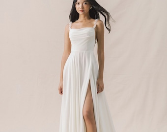 May include: A white sleeveless wedding dress with a cowl neck and a thigh-high slit. The dress has a delicate floral detail on the straps.