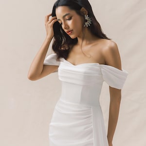 May include: A white off-the-shoulder dress with a fitted bodice and a flowing skirt. The dress has a side slit and a ruched detail on the skirt.