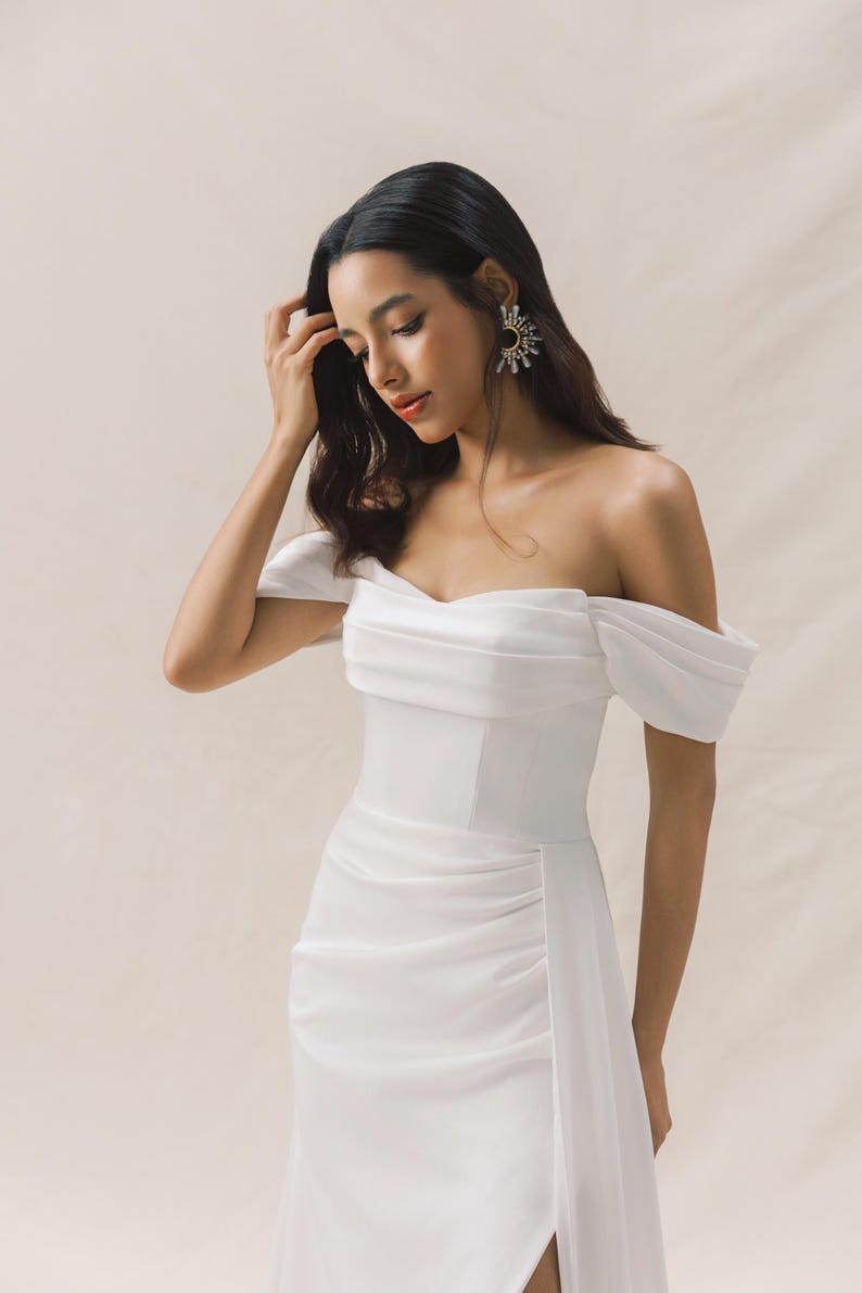 May include: A white off-the-shoulder dress with a fitted bodice and a flowing skirt. The dress has a side slit and a ruched detail on the skirt.