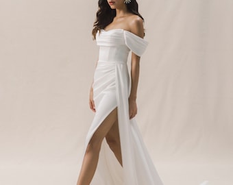 May include: A white off-the-shoulder gown with a fitted bodice and a flowing skirt. The skirt has a high slit on the left side and a train.