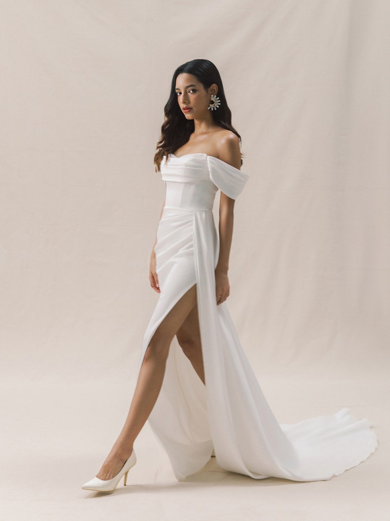 Bride in an off-shoulder satin wedding dress with a fitted bodice, thigh-high slit, and subtle train. Elegant and modern, with statement earrings and heels.