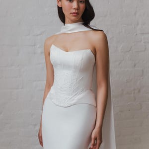 May include: A white wedding dress with a unique design. The dress features a strapless, structured bodice with intricate embroidered detailing. A matching white cape drapes over one shoulder, adding a flowing element. The skirt is a simple, sleek design. The model is standing in front of a white brick wall.