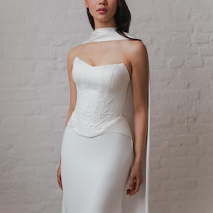 May include: A white strapless wedding dress with a unique design. The bodice features intricate embroidered detailing, and the skirt is a simple, flowing design. A matching white cape is draped over the shoulders, adding a touch of elegance. The dress is displayed against a white brick wall.