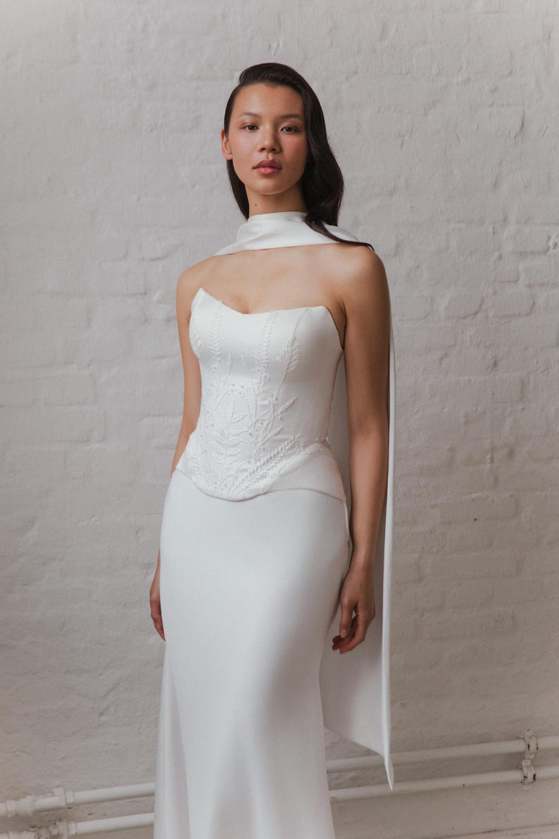 May include: A white strapless wedding dress with a unique design. The bodice features intricate embroidered detailing, and the skirt is a simple, flowing design. A matching white cape is draped over the shoulders, adding a touch of elegance. The dress is displayed against a white brick wall.