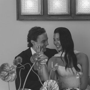 May include: A black and white photograph captures a couple at a table, smiling and looking at each other. The woman wears a strapless dress, and the man is in a suit. The table is set with plates, glasses, and floral arrangements. A wall with square, illuminated panels is in the background.