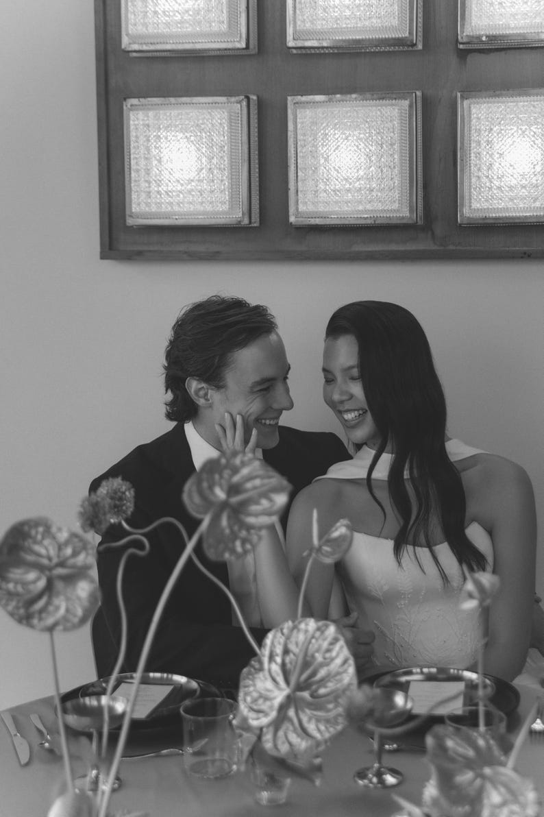 May include: A black and white photograph captures a couple at a table, smiling and looking at each other. The woman wears a strapless dress, and the man is in a suit. The table is set with plates, glasses, and floral arrangements. A wall with square, illuminated panels is in the background.