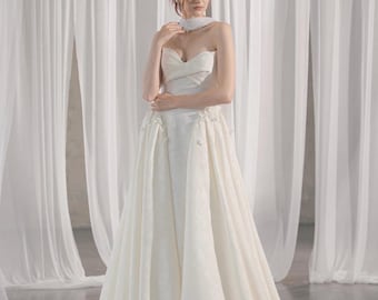 Puede incluir: Vestido de novia marfil sin tirantes con escote corazón y falda amplia. El vestido presenta detalles florales y un choker a juego. La modelo posa frente a cortinas blancas.