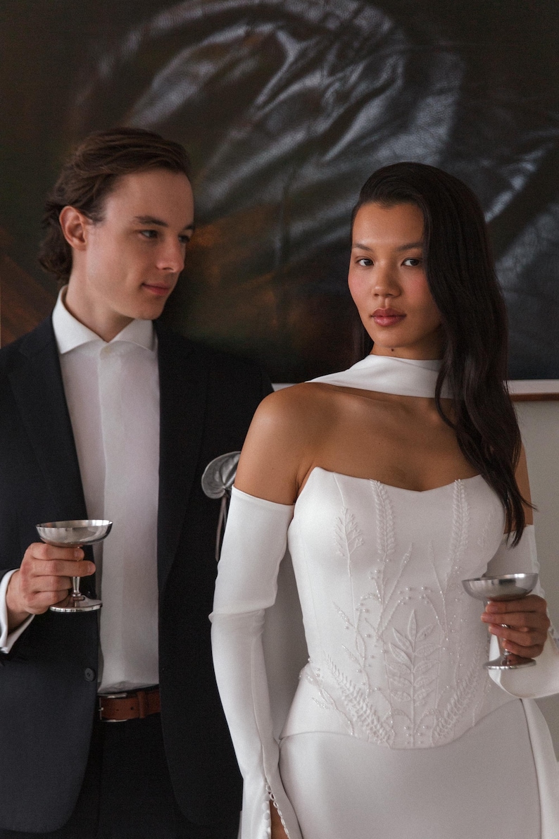 May include: A formal ensemble featuring a white strapless gown with intricate leaf-like embroidery on the bodice, paired with long white gloves and a matching scarf. A black suit with a white dress shirt and a silver champagne glass is held by a person standing next to the gown.