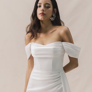 Bride in an off-shoulder satin wedding dress with a fitted bodice, thigh-high slit, and subtle train. Elegant and modern, with statement earrings and heels.