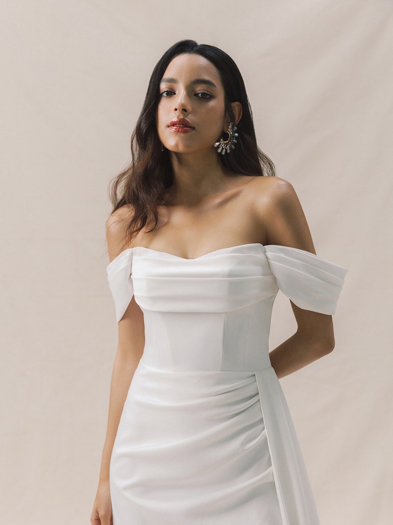 Bride in an off-shoulder satin wedding dress with a fitted bodice, thigh-high slit, and subtle train. Elegant and modern, with statement earrings and heels.