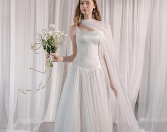 May include: A white wedding dress with a flowing skirt and a fitted bodice. The dress features delicate embroidery and is paired with a sheer, draped shawl. The bride holds a bouquet of white flowers. The background is a white curtain.
