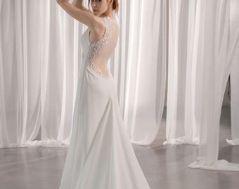 May include: A white wedding dress with a sheer, embellished back and a flowing skirt. The sleeveless gown features a scoop neckline and a subtle train. The model is posing in front of white draped fabric.
