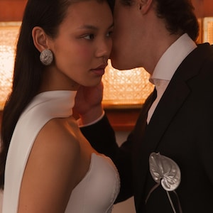 May include: A woman in a white wedding dress with a high neckline and sheer sleeves. She wears a large, round, silver earring. A man in a black suit with a white collared shirt leans in, whispering near her ear. A silver leaf-shaped boutonniere is pinned to his lapel.