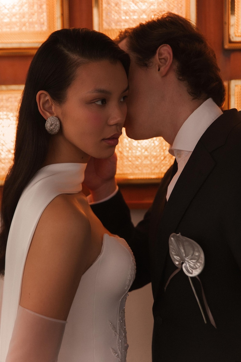 May include: A woman in a white wedding dress with a high neckline and sheer sleeves. She wears a large, round, silver earring. A man in a black suit with a white collared shirt leans in, whispering near her ear. A silver leaf-shaped boutonniere is pinned to his lapel.