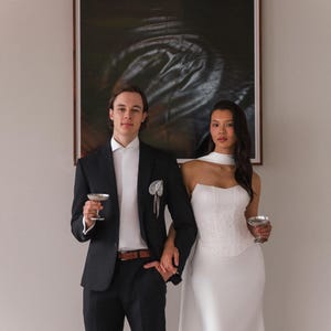 May include: A couple stands holding champagne glasses in front of a large abstract painting. The person on the left wears a black suit with a white shirt and a pocket square. The person on the right wears a white, strapless dress with a fitted bodice and a flowing skirt.