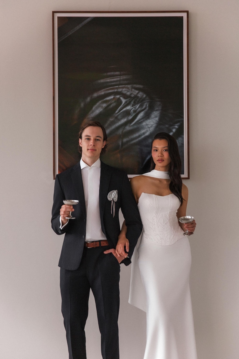 May include: A couple stands holding champagne glasses in front of a large abstract painting. The person on the left wears a black suit with a white shirt and a pocket square. The person on the right wears a white, strapless dress with a fitted bodice and a flowing skirt.