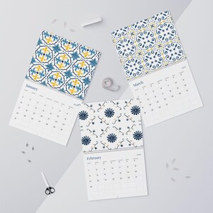 Our 2027 Azulejos  wall calendar featuring January, February, and March pages. Each page showcases a beautiful watercolor tile pattern in shades of blue and yellow. The calendars are spiral-bound and laid flat with scattered paperclips and scissors.