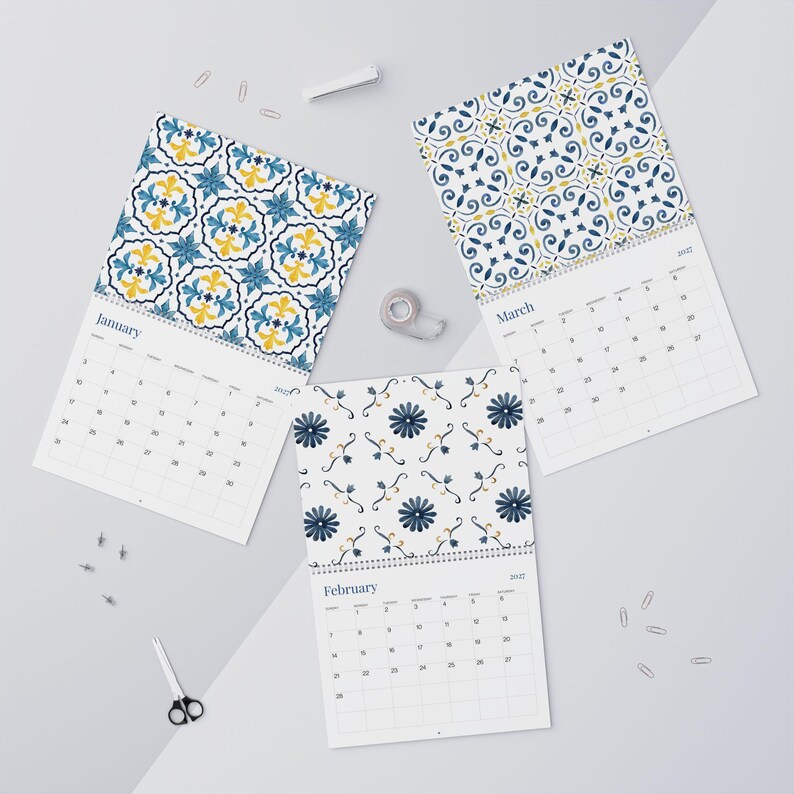 Our 2027 Azulejos  wall calendar featuring January, February, and March pages. Each page showcases a beautiful watercolor tile pattern in shades of blue and yellow. The calendars are spiral-bound and laid flat with scattered paperclips and scissors.
