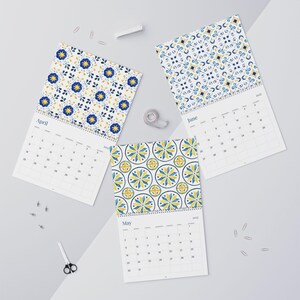 This flat lay image showcases three open 2027 Azulejos calendars with wire binding. Each calendar features a decorative top with a blue and yellow tile-inspired floral pattern. The calendars display the months of April, May, and June. Office supplies like paper clips, tape, a stapler, and scissors are scattered around the calendars on a light gray background.