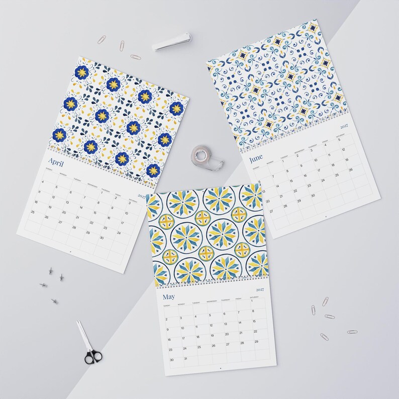 This flat lay image showcases three open 2027 Azulejos calendars with wire binding. Each calendar features a decorative top with a blue and yellow tile-inspired floral pattern. The calendars display the months of April, May, and June. Office supplies like paper clips, tape, a stapler, and scissors are scattered around the calendars on a light gray background.