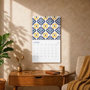 Our 2027 Azulejos wall calendar opened to October 2027, featuring a vibrant geometric floral pattern in navy blue, yellow, and red on a white background. The calendar grid below shows the month and dates. The page is hung on a textured, neutral-toned wall with soft, dappled shadows cast from a nearby window. A mid-century modern wooden desk sits below, adorned with a brass vase holding dried flowers, a decorative key, a leather-bound journal, and a ceramic cup.