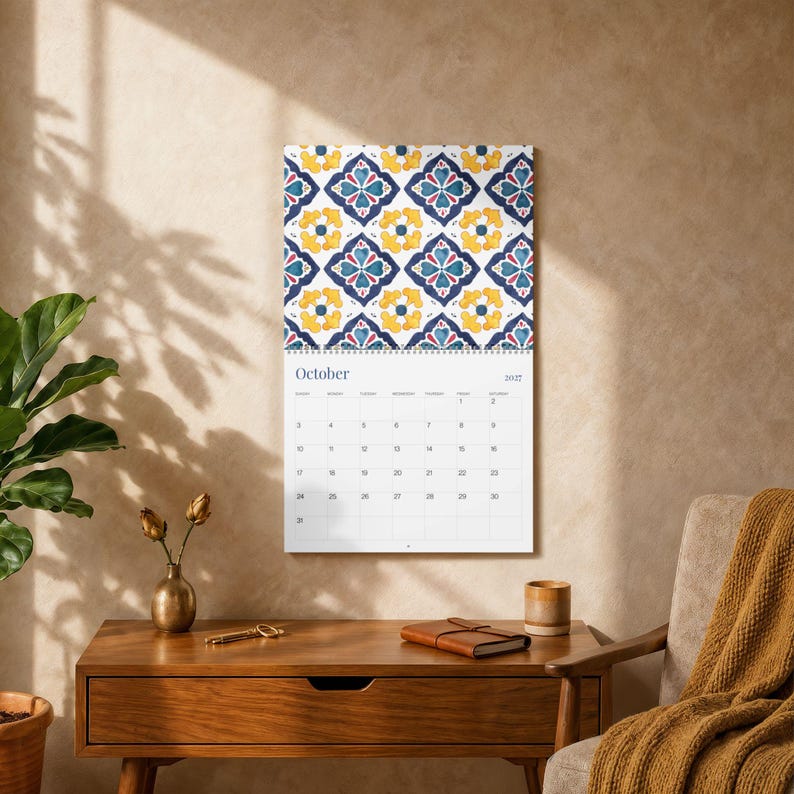 Our 2027 Azulejos wall calendar opened to October 2027, featuring a vibrant geometric floral pattern in navy blue, yellow, and red on a white background. The calendar grid below shows the month and dates. The page is hung on a textured, neutral-toned wall with soft, dappled shadows cast from a nearby window. A mid-century modern wooden desk sits below, adorned with a brass vase holding dried flowers, a decorative key, a leather-bound journal, and a ceramic cup.