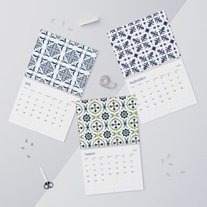 Our beautiful 2027 Azulejos wall calendar featuring vibrant, patterned pages for July, August, and September. The designs include intricate, repeating floral motifs in shades of deep blue on white, and a colorful tile pattern with green, blue, and yellow accents. The calendars display clearly marked dates and days of the week, arranged for easy viewing.