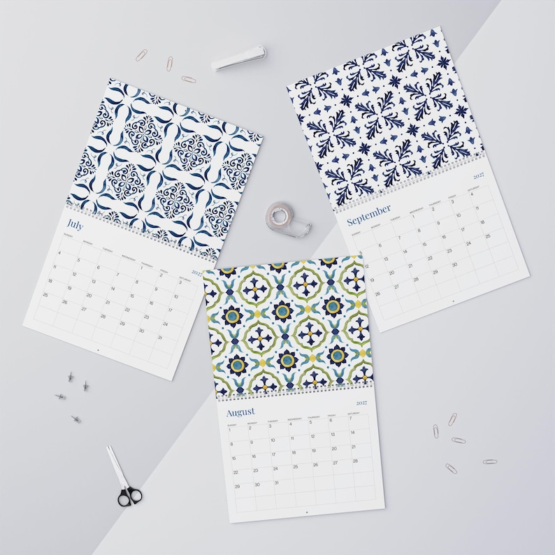 Our beautiful 2027 Azulejos wall calendar featuring vibrant, patterned pages for July, August, and September. The designs include intricate, repeating floral motifs in shades of deep blue on white, and a colorful tile pattern with green, blue, and yellow accents. The calendars display clearly marked dates and days of the week, arranged for easy viewing.