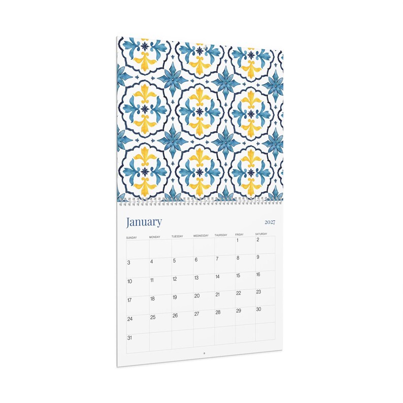 A close-up angled image of our 2027 Azulejos wall calendar opened to January, featuring a vibrant blue and yellow floral tile pattern at the top. The calendar grid below has days of the week and dates clearly displayed.