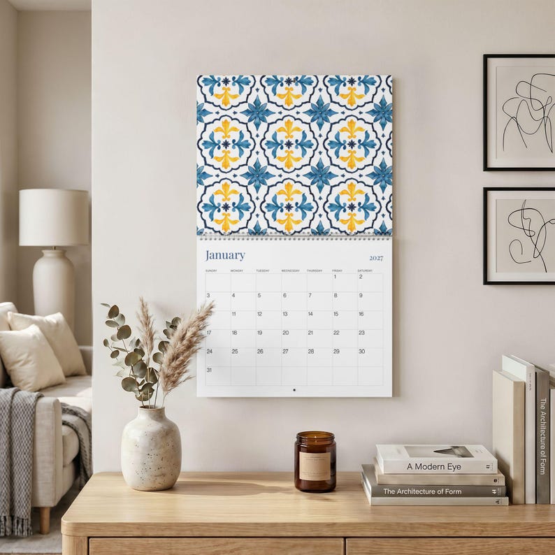 Our 2027 Azulejos Mediterranean inspired wall calendar opened to January 2027 featuring a blue and yellow floral tile pattern at the top hangs on a neutral wall. Below, a modern desk holds a speckled ceramic vase with dried eucalyptus and pampas grass, a lit amber candle, and a stack of minimalist books.