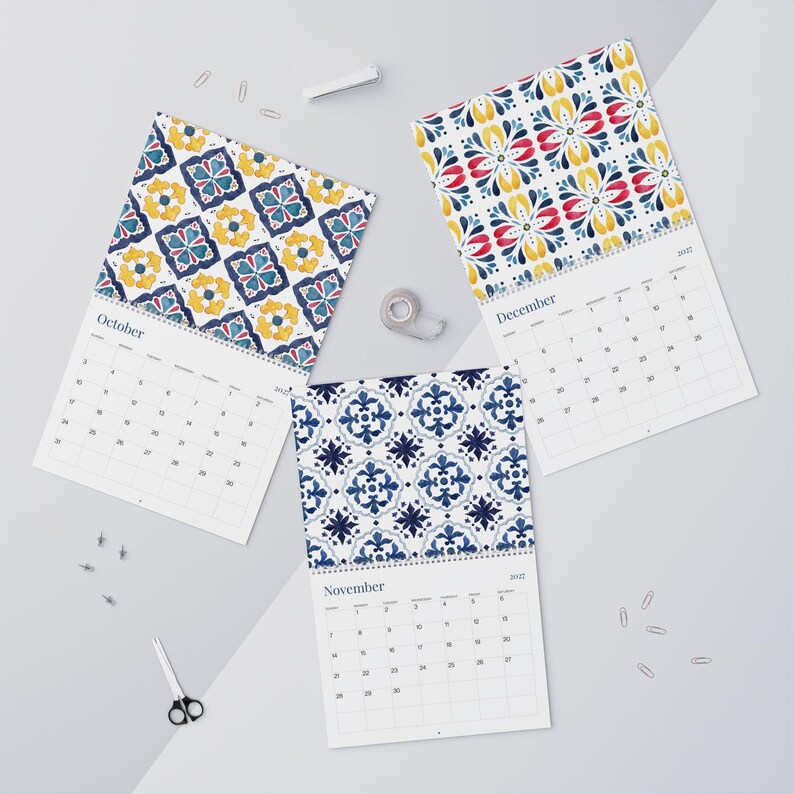 Our 2027 Azulejos wall calendar showcasing October, November, and December feature beautiful watercolor tile patterns. October has a yellow, blue, and red floral design. November displays a rich blue tile motif. December is adorned with a vibrant pattern of red, blue, and yellow floral elements. The calendars are displayed flat on a light gray background with paper clips and scissors.
