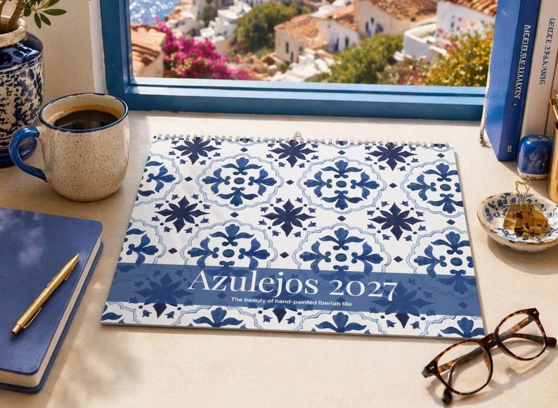 Our 2027 Mediterranean inspired wall calendar featuring "Azulejos" in blue and white tile patterns. It sits on a desk next to a blue notebook, a gold pen, a speckled mug of coffee, and eyeglasses. A scenic view of a white Mediterranean village overlooking the blue sea is visible through a blue-framed window.