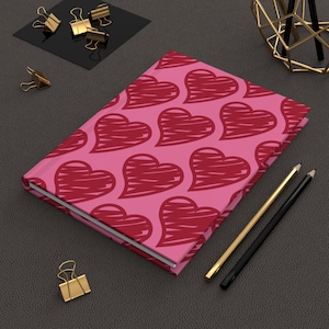 May include: A pink journal with a pattern of red sketched hearts. Gold and black pencils and gold binder clips are arranged around the journal on a dark gray surface. A modern desk accessory.
