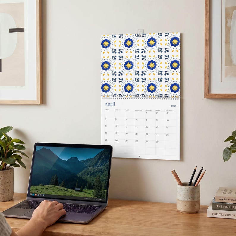Our 2027 Azulejos Wall calendar opened to April 2027 with a vibrant floral tile pattern in blue, yellow, and navy on a white background. Calendar dates are clearly visible with a clean, modern font. A laptop, potted plant, and desk accessories complete the cozy, productive workspace.