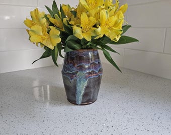 May include: A ceramic vase with a dark brown and blue glaze holds a bouquet of yellow flowers with green leaves. The vase has a unique, textured appearance and sits on a light-colored countertop. The flowers are in full bloom.